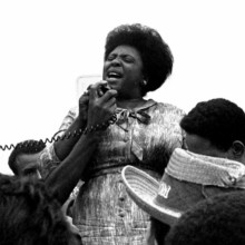 Black and white photograph of Fannie Lou Hammer. Three quarter photo of Black woman holding and speaking into a speaker microphone. The heads of other people can be seen around her waist.