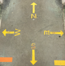 Image of a yellow compass painted on the floor of the Hatcher Library’s North Stacks, with arrows pointing outward to the four directions. 