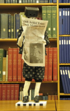 Figurine reading a newspaper, with a box for a head. 