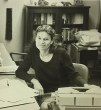 Photograph: Portrait of Eleanor Burke Leacock sitting at a desk, with a book case visible behind her. 