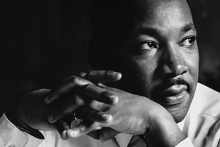 Black and white photograph of Dr. Martin Luther King. Black male seen from shoulders up, looking off to the left with hands folded in front of him. 