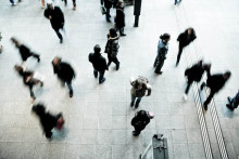 Photo taken above a busy crowd. Some figures are actively walking and their figure is blurred from their movement. Others figures are clear and sharp and they are standing, talking to or watching other people in the crowd.