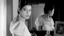 Black and white film still showing a Black woman from the chest up, looking directly at the camera. Also within the frame is the back of her head as shown in a mirror behind her. 